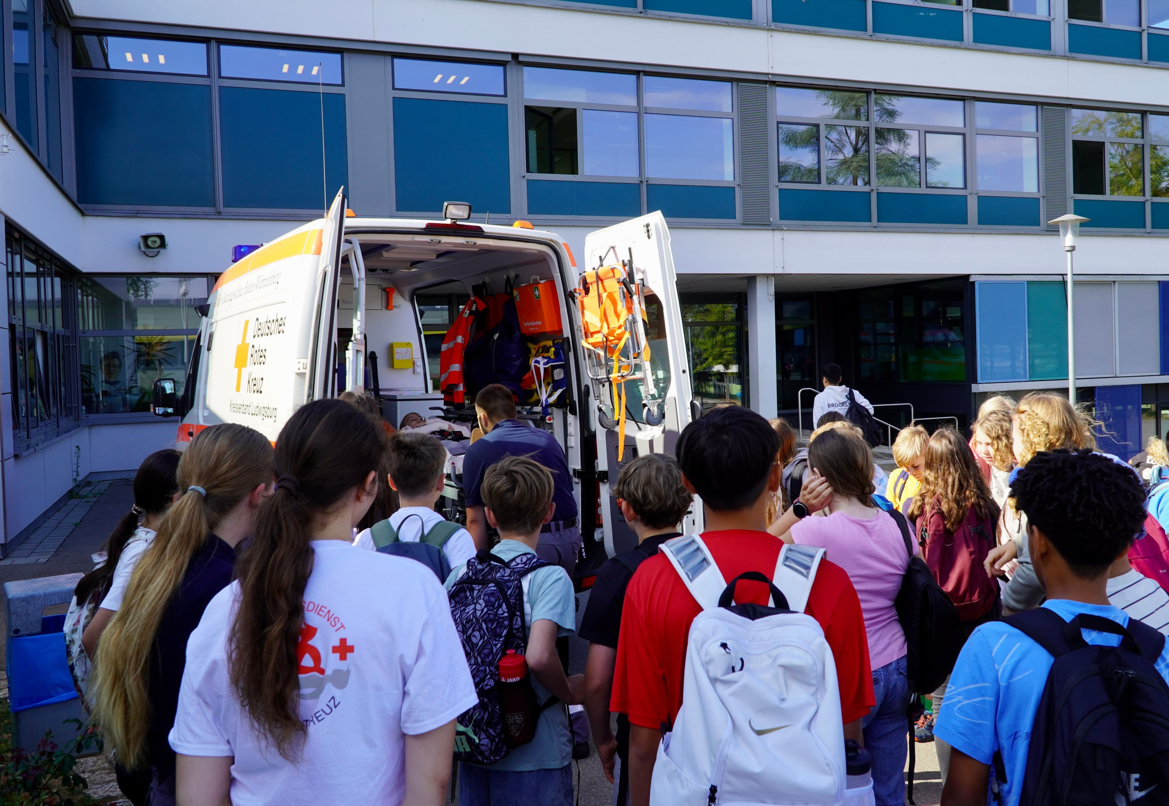 Students look into an open German Red Cross ambulance parked in front of a building.