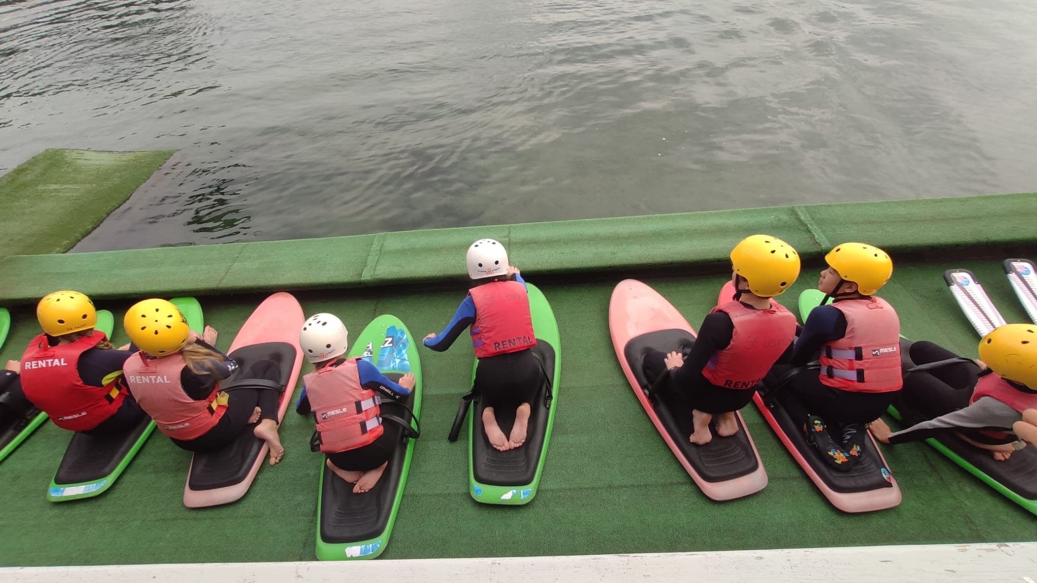 Eine Gruppe von Kindern sniet auf Wasserboards am Ufer eines Sees
