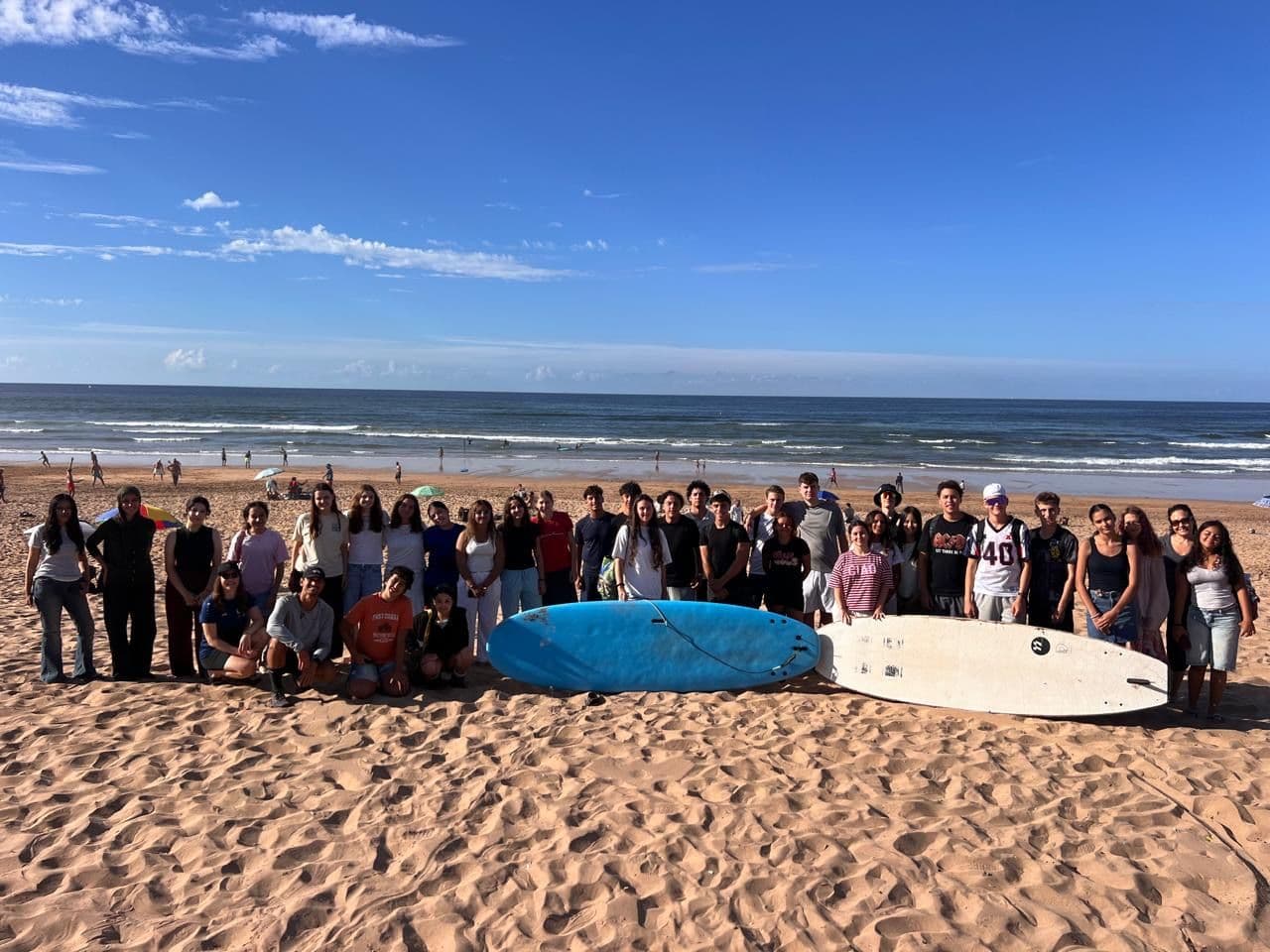 A large group of young people poses on a sandy beach with two surfboards, the ocean in the background.