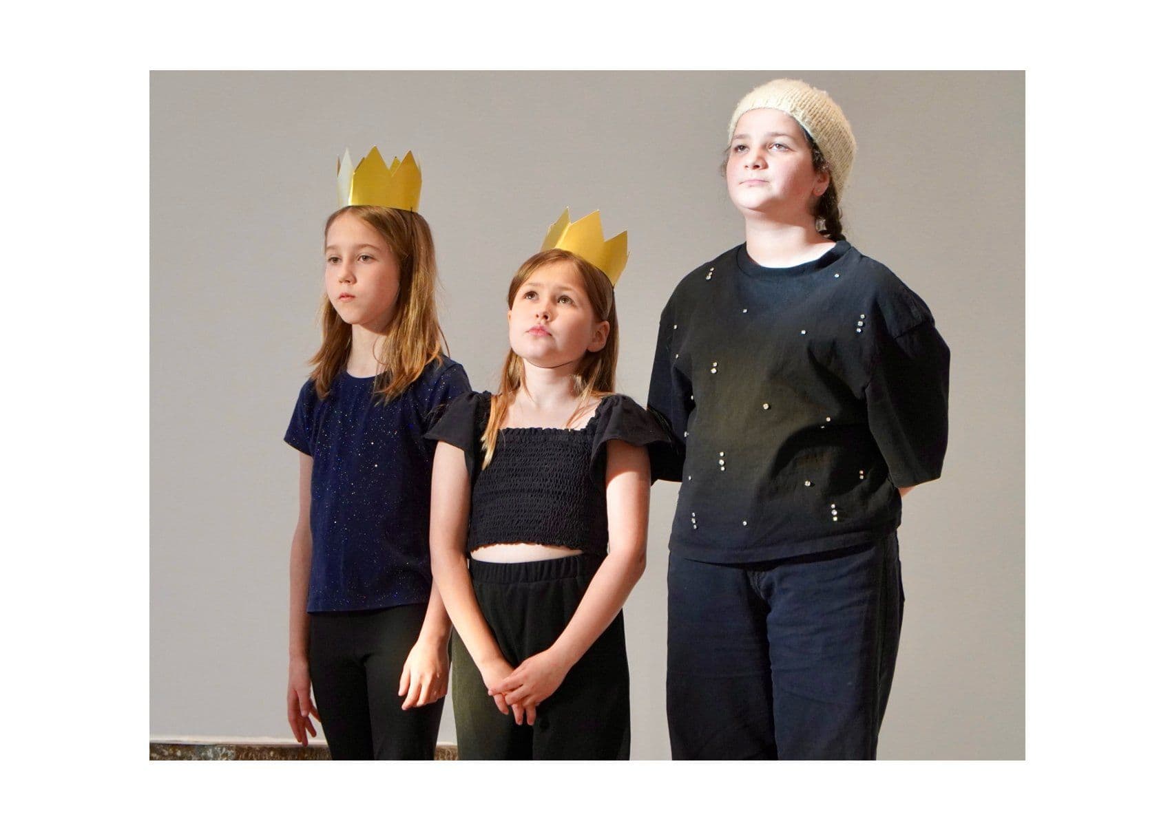Three children, two wearing gold paper crowns and one a light beanie, stand looking upward.