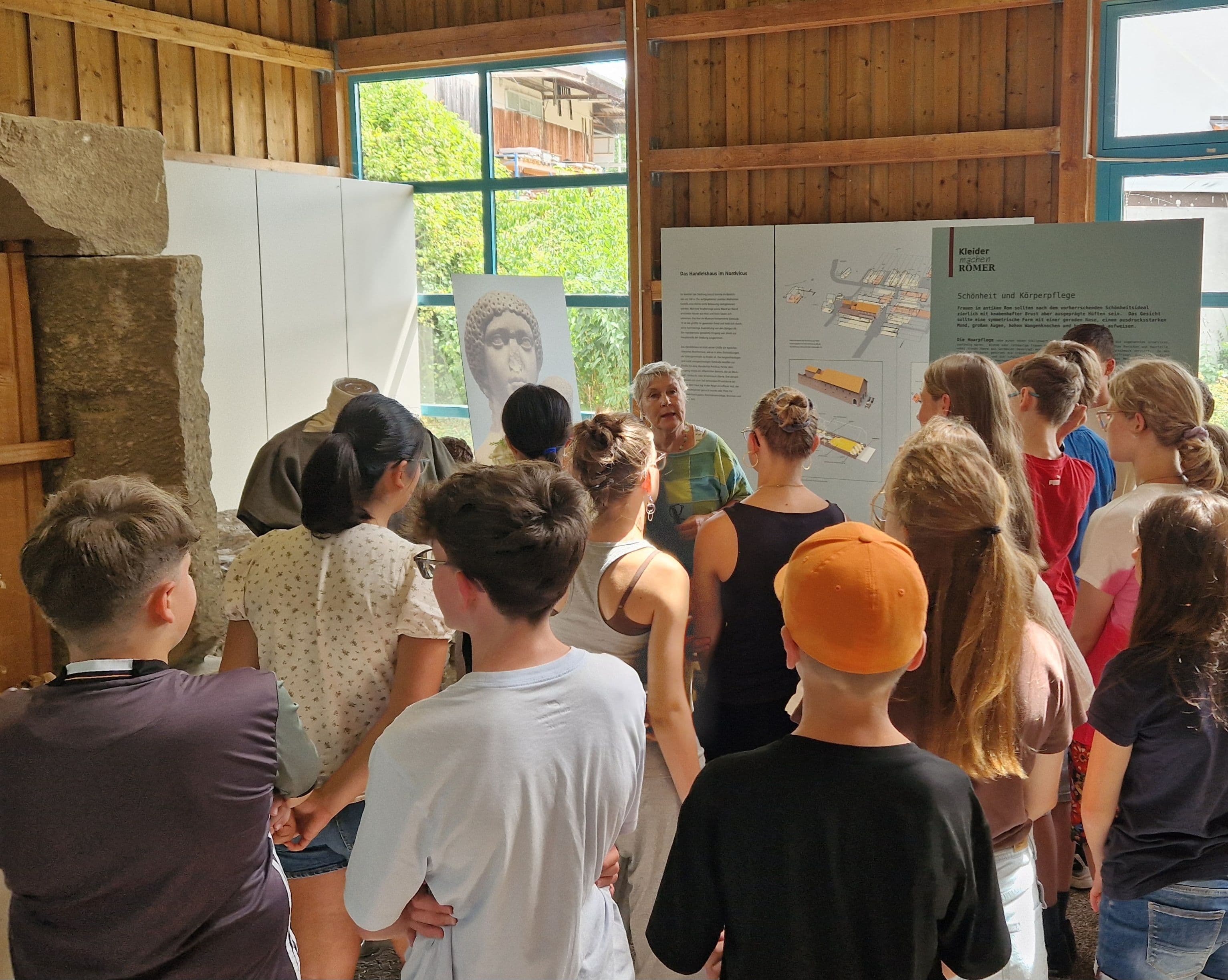 An older woman guides a group of young people through a rustic museum exhibit featuring a large bust, stone blocks, and informational displays.