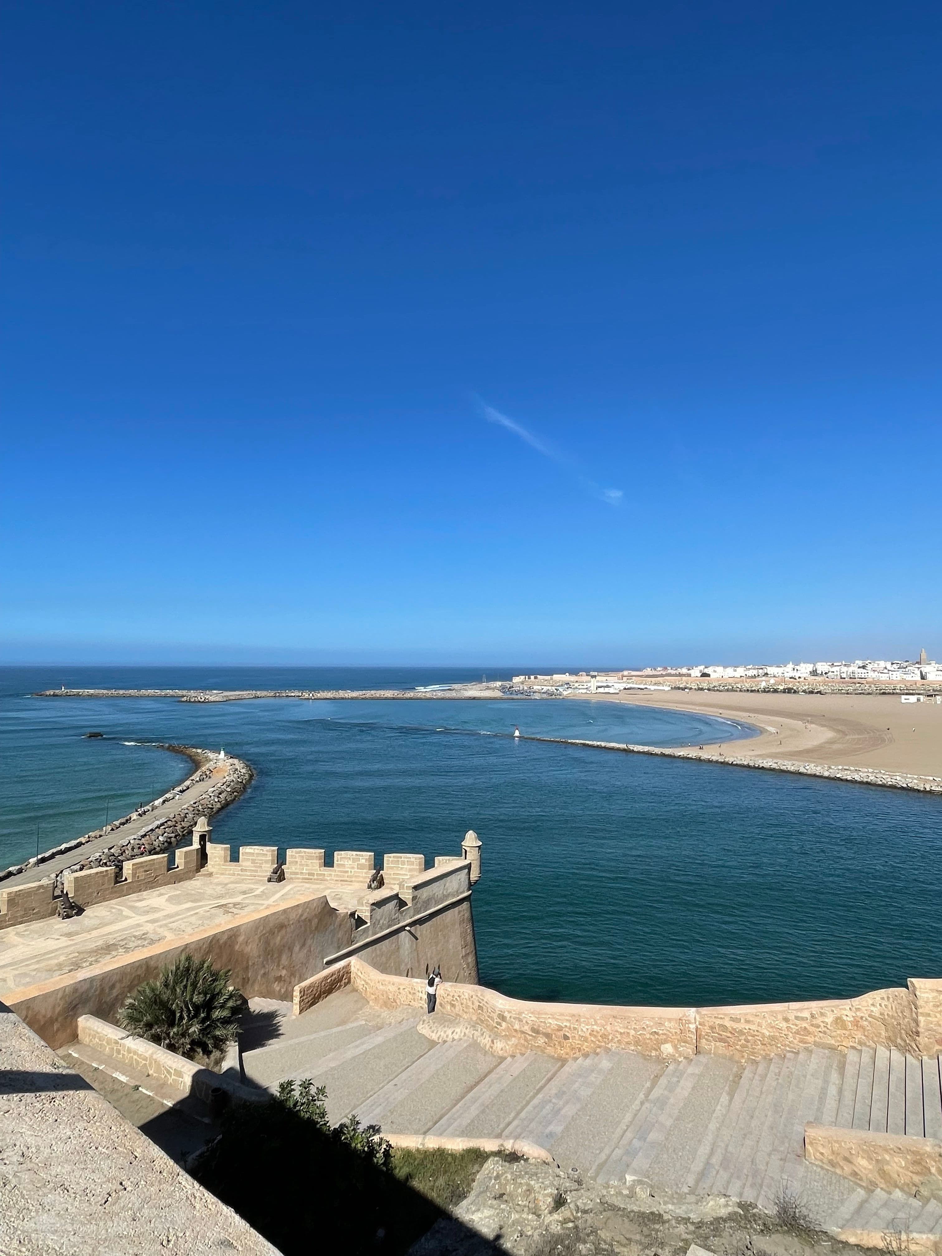 A historic stone fortress overlooks a river estuary flowing into the ocean, with a beach and town visible in the distance under a clear blue sky.