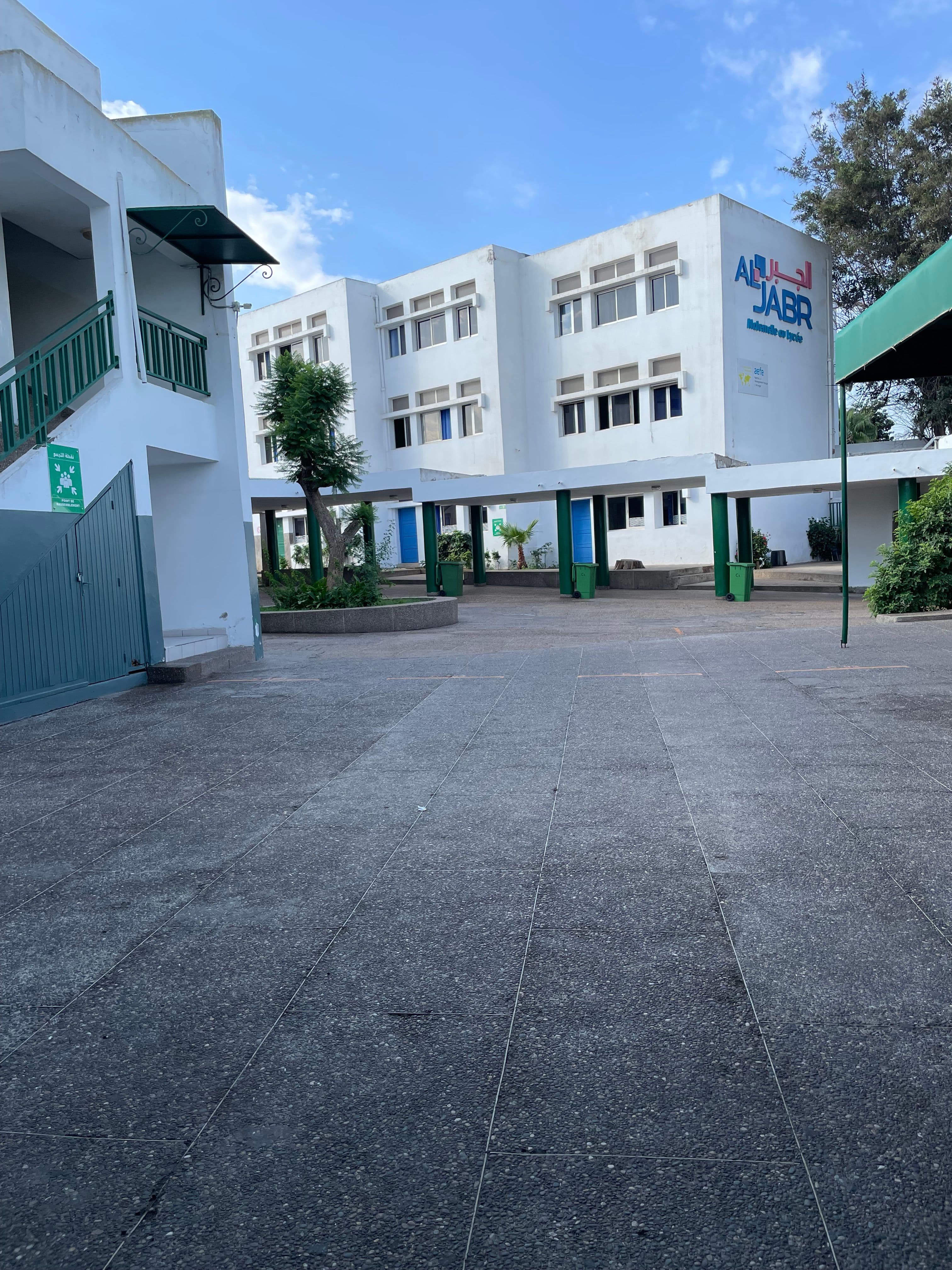 Exterior view of the white AL JABR school building, a paved courtyard, green stairs, and covered walkways under a blue sky.