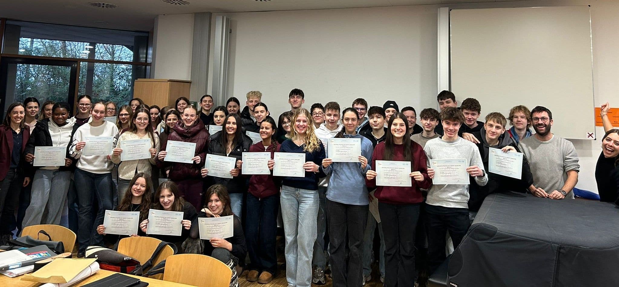 A large group of smiling students and adults holds up certificates in a classroom.