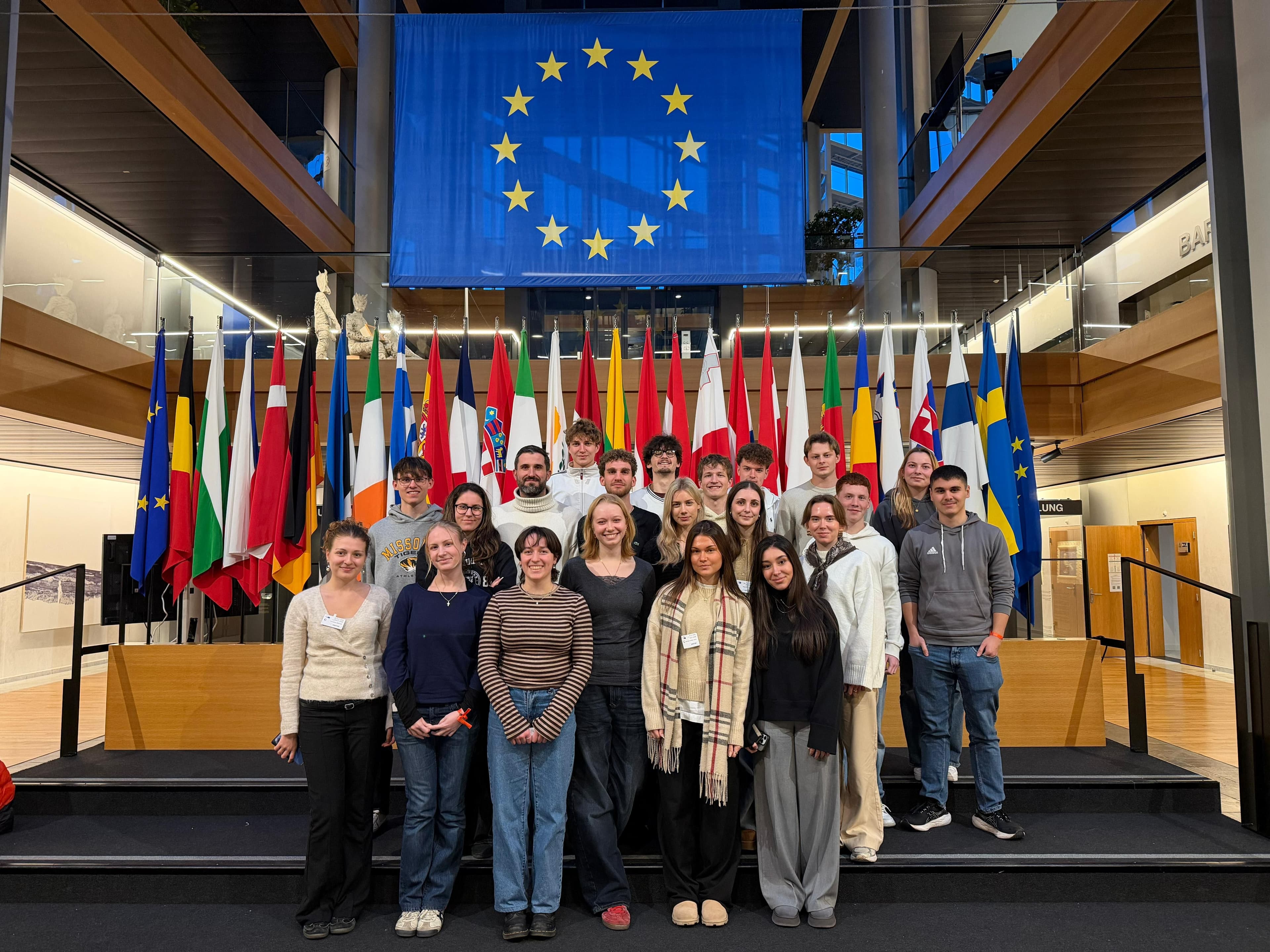 A group of young adults stands on steps in front of a large European Union flag and numerous national flags.