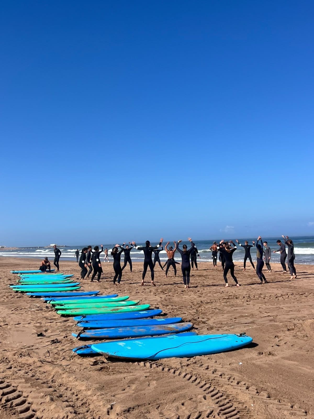 A group of people in wetsuits stretch on a sunny beach, with rows of colorful surfboards in the sand and the ocean in the background.