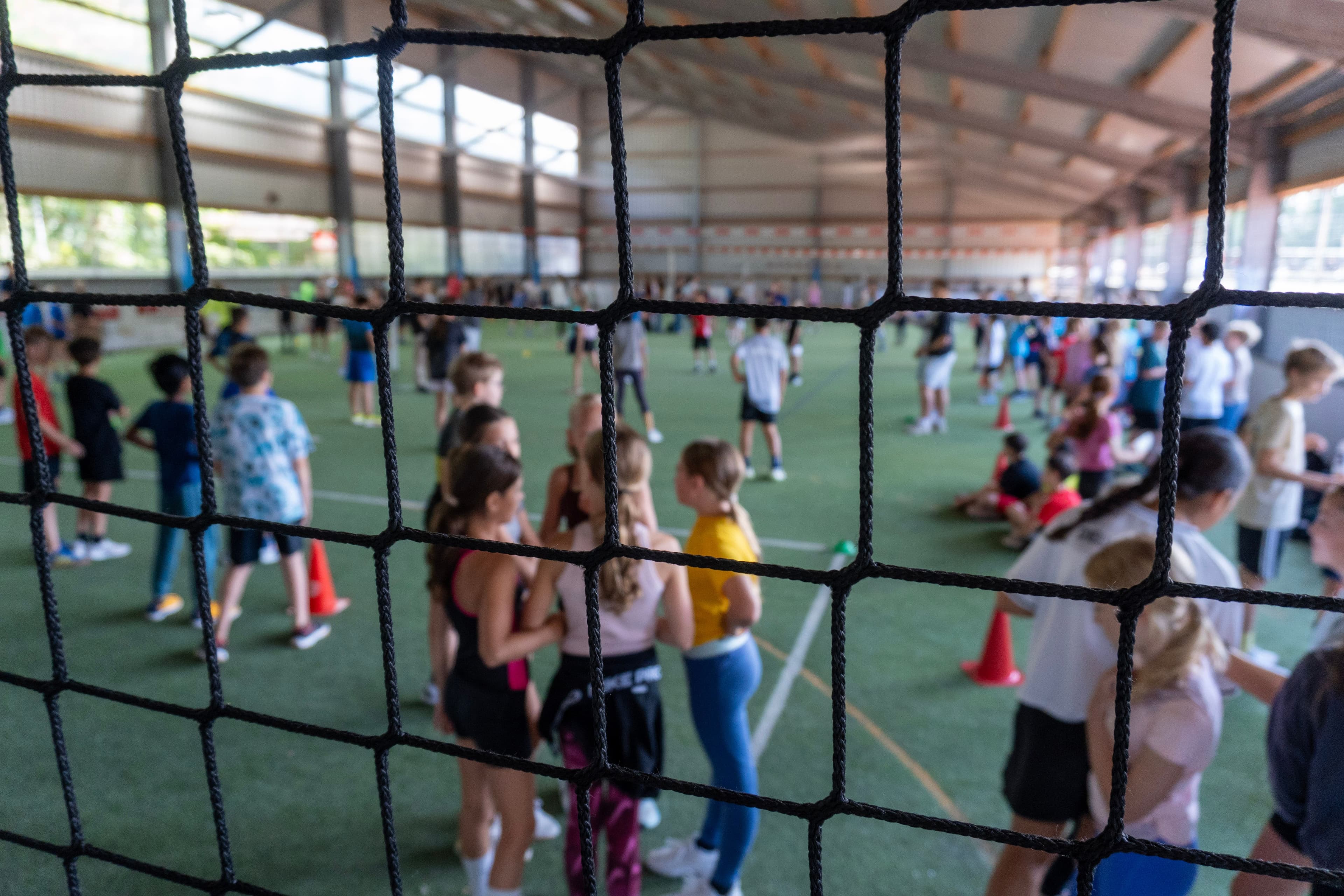 Blurred children in an indoor sports facility, viewed through a sharp black net.