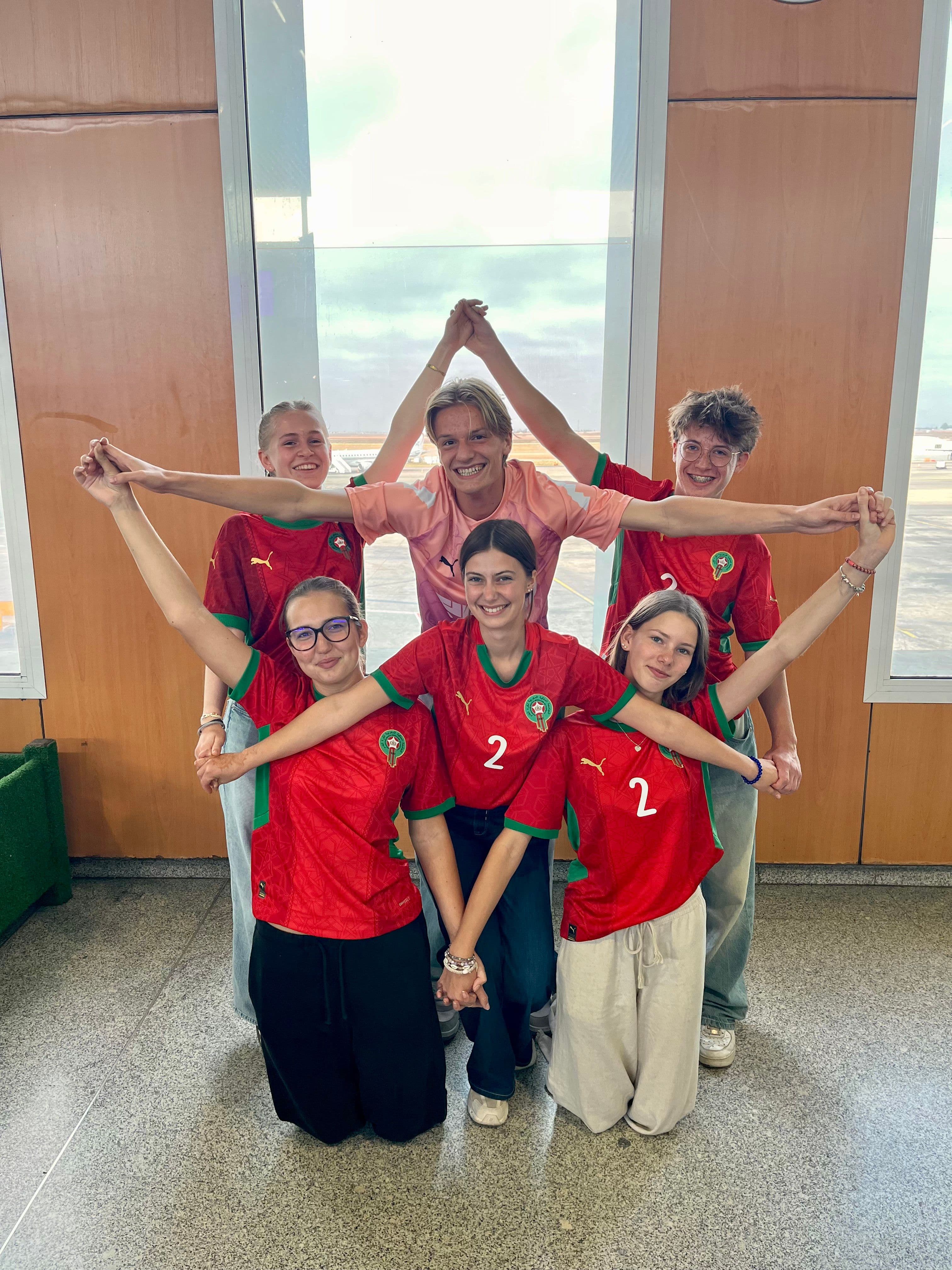 Six smiling young people, mostly in red Moroccan soccer jerseys, form a large W with linked arms in an airport terminal.