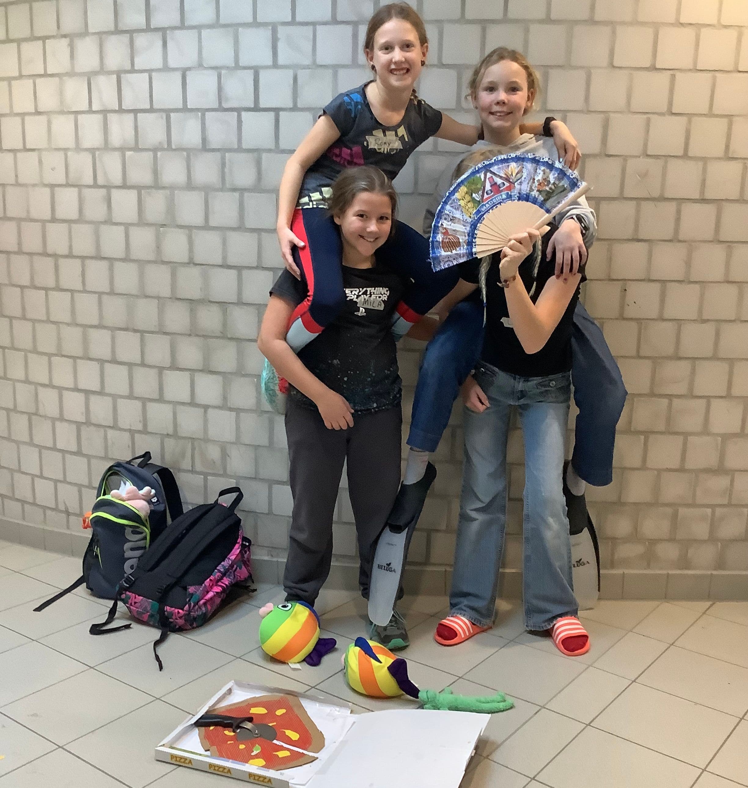 Four smiling girls in two piggyback pairs, one holding a fan, two wearing flippers, with backpacks and toys on the floor.