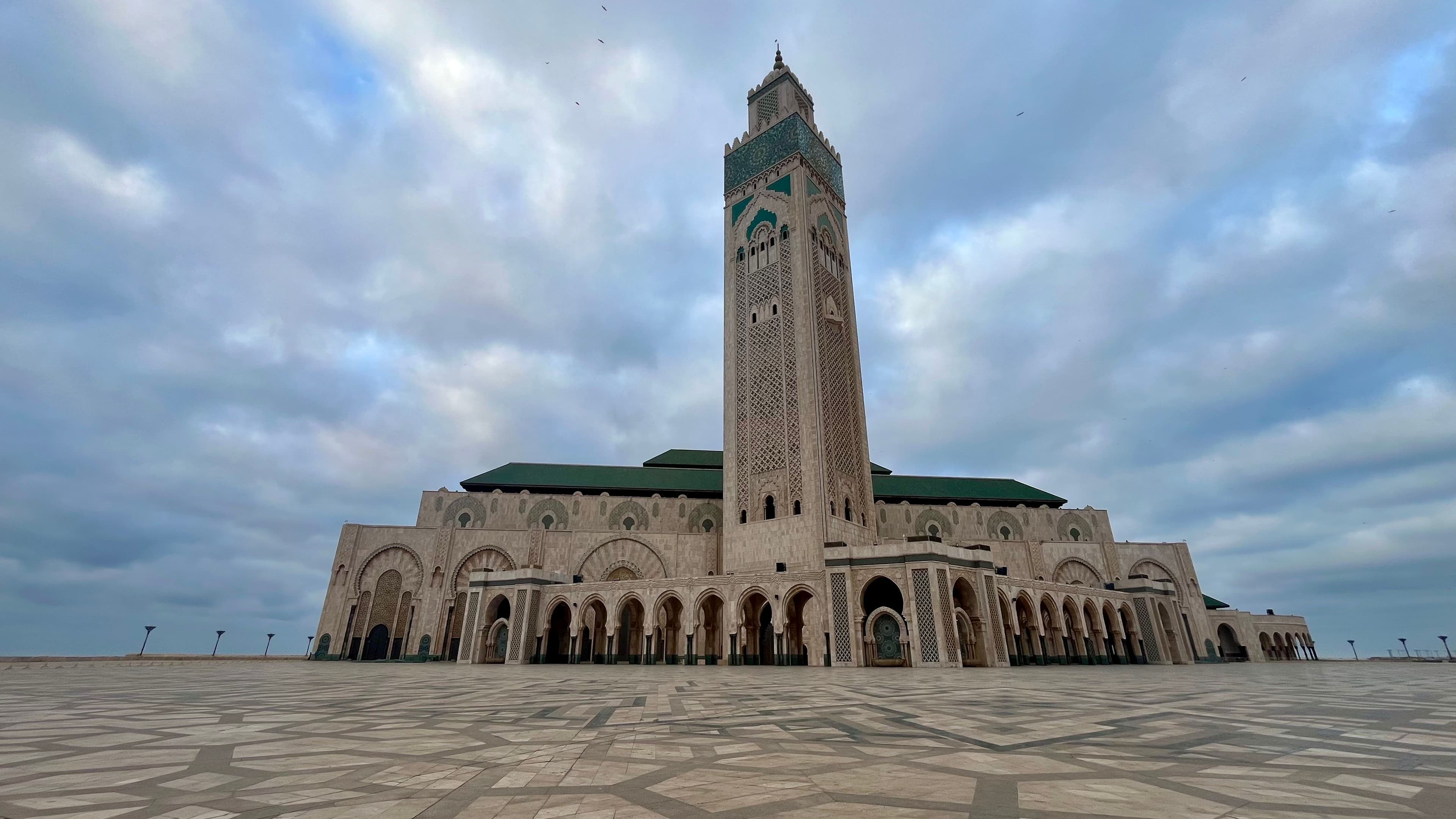 Hassan II Mosque with a tall minaret, viewed from a large, patterned plaza under a cloudy sky.