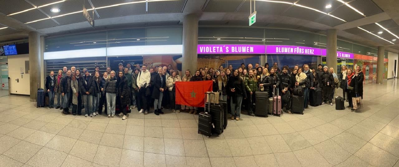 A large group of people with luggage holding a Moroccan flag in an airport terminal.
