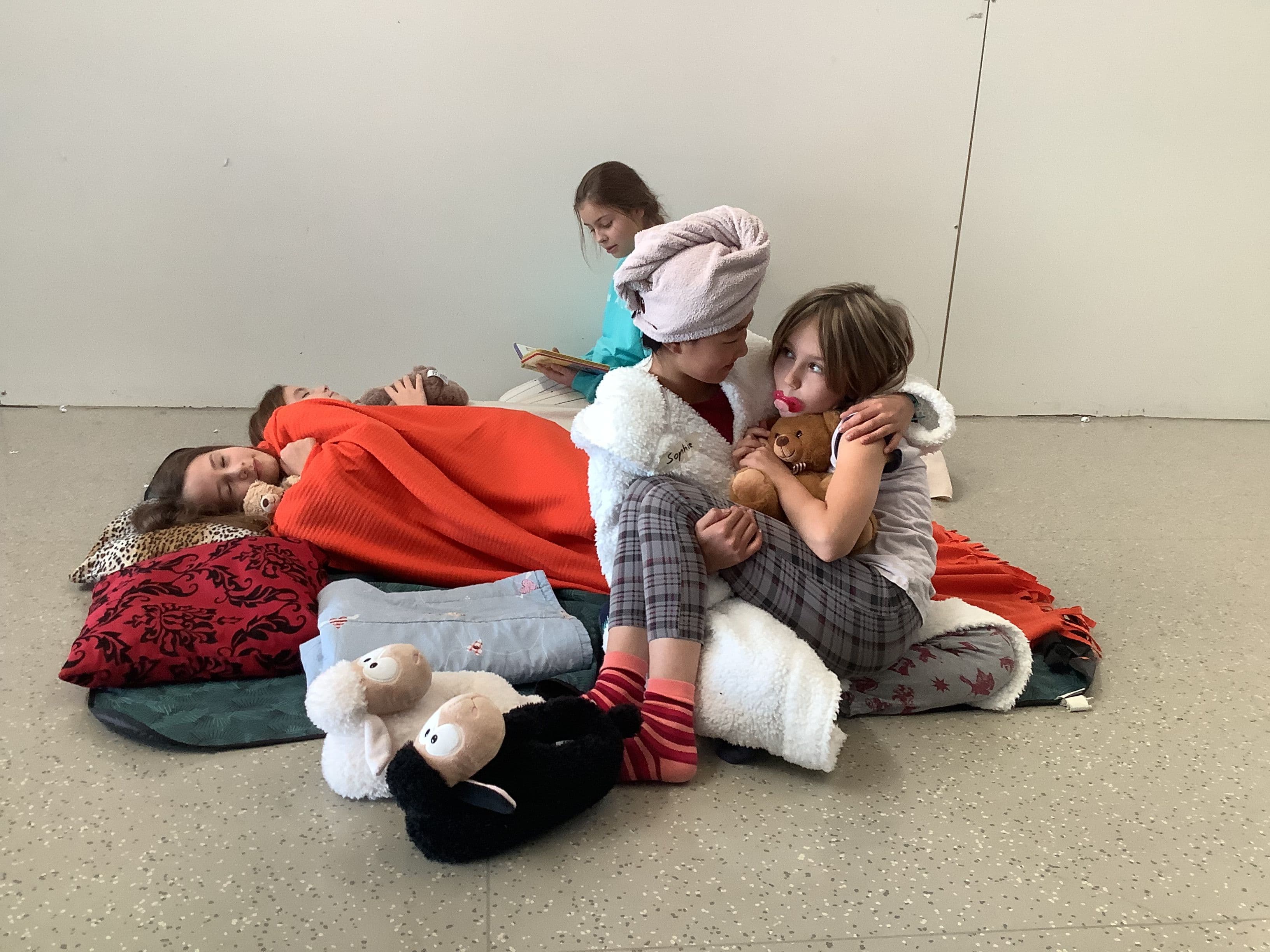 Several girls are having a sleepover on the floor with blankets and pillows. One girl sleeps, another reads, and two girls hug in the foreground, one wearing a towel and the other holding a teddy bear with a pacifier.