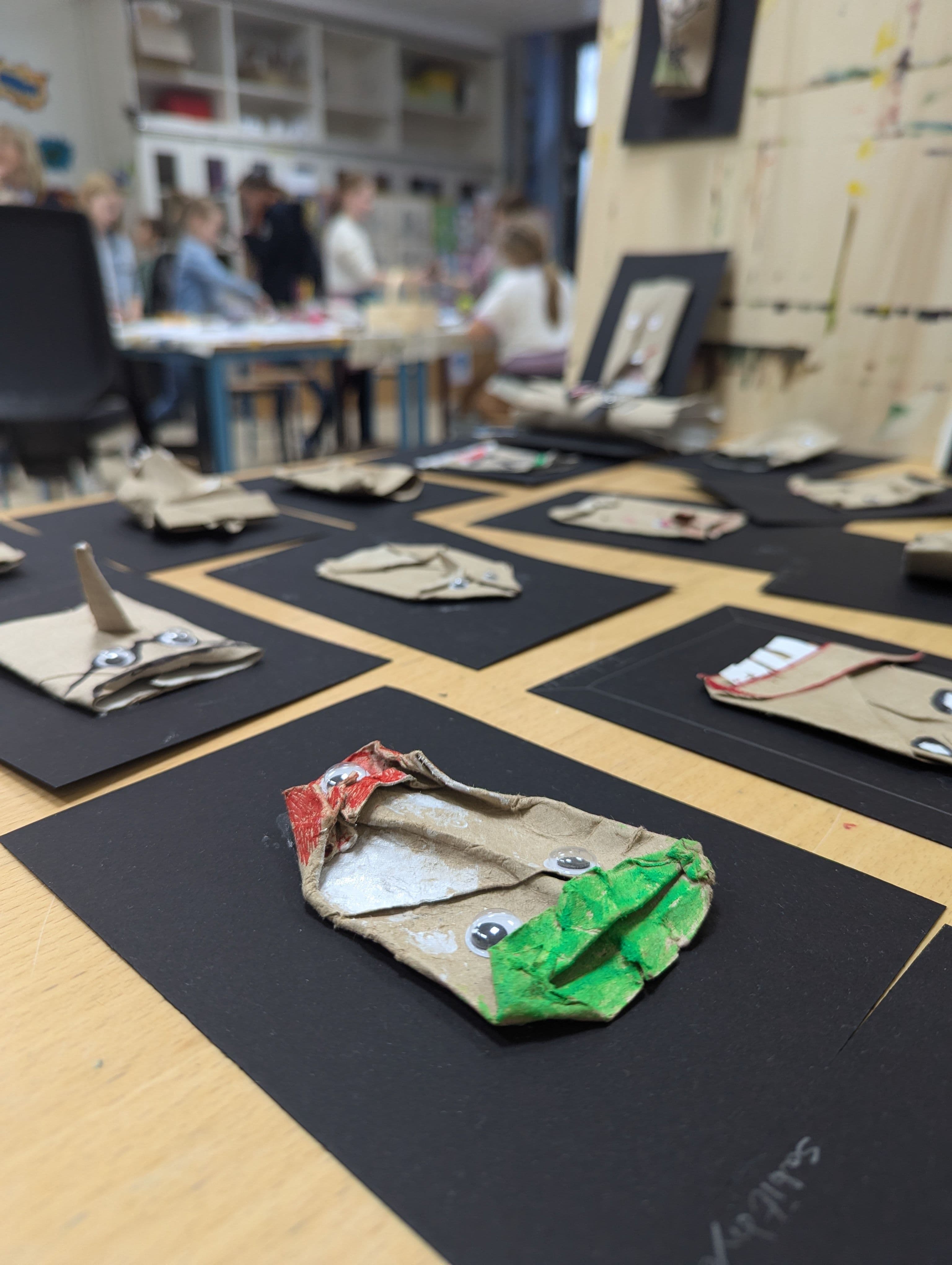 gefaltete Papprollen, die zu Gesichtern umgestaltet wurden , liegen auf einem Tisch. Im Hintergrund ist eine Kunstraum mit unscharfen Menschen abgebildet.