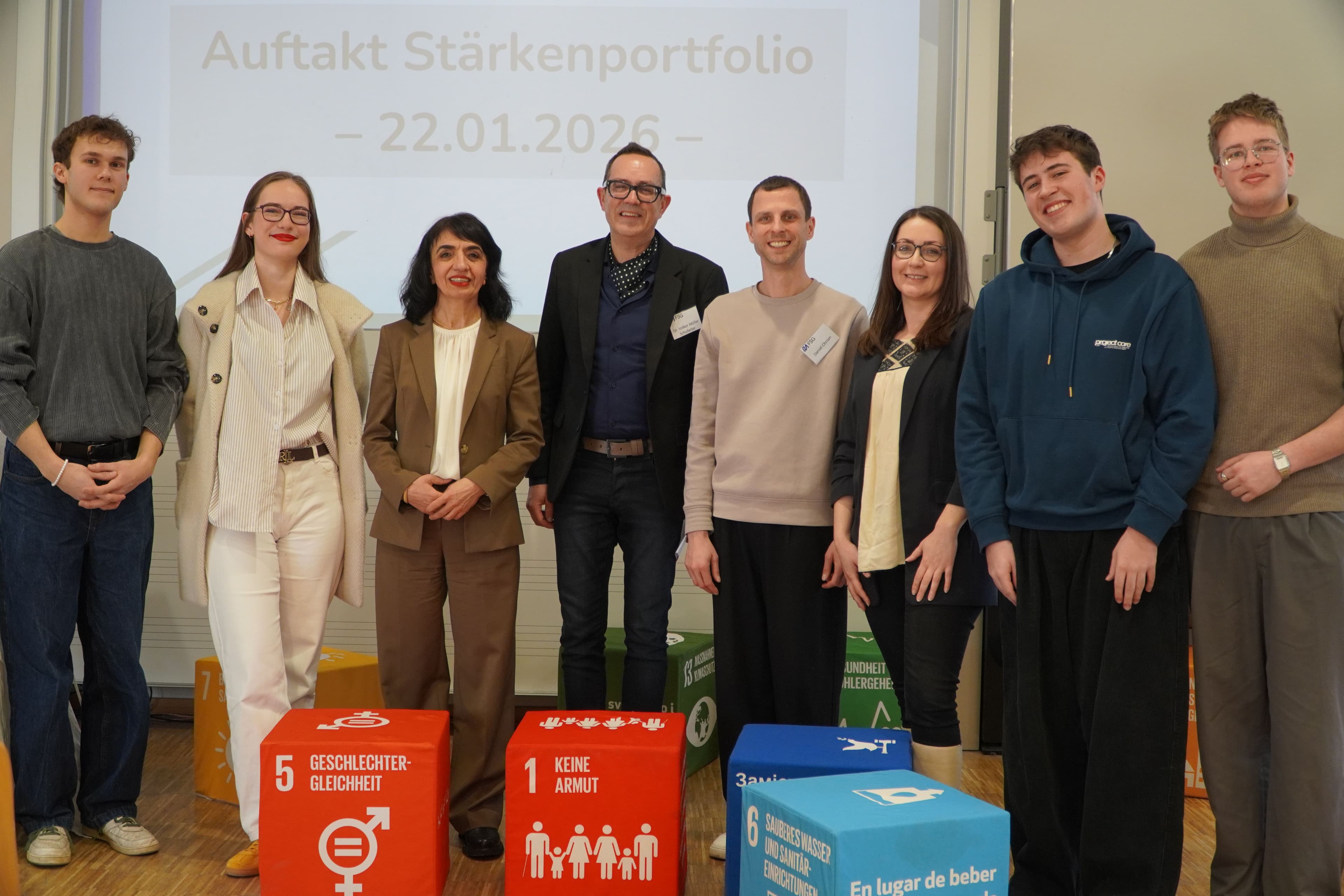 Eight people stand smiling in front of a projector screen and colorful cubes illustrating UN Sustainable Development Goals.