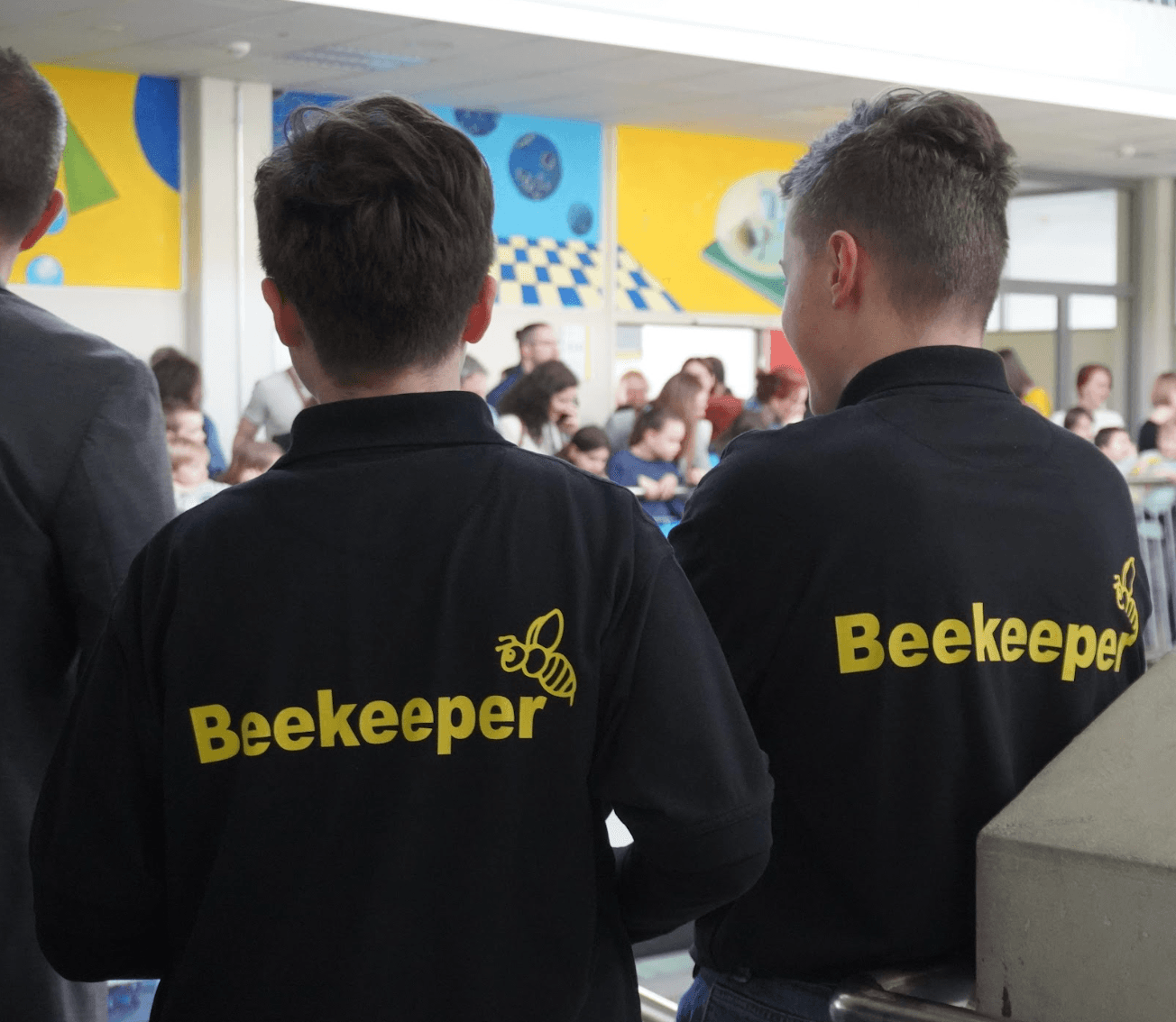 Two people from behind wear black shirts with "Beekeeper" and a bee logo, facing a crowd in a room with colorful murals.
