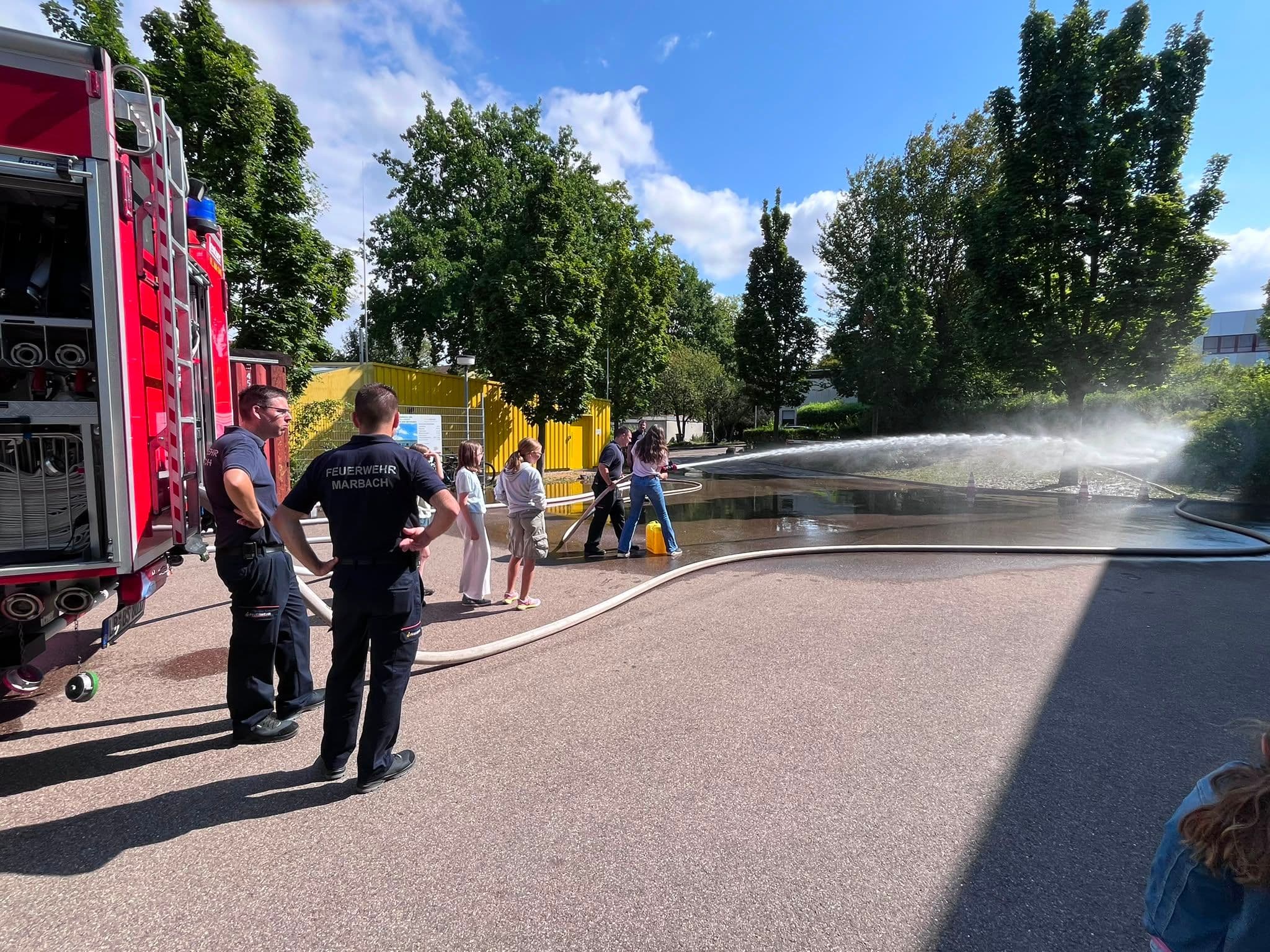 Firefighters demonstrate a fire hose to children from a fire truck on a sunny day.