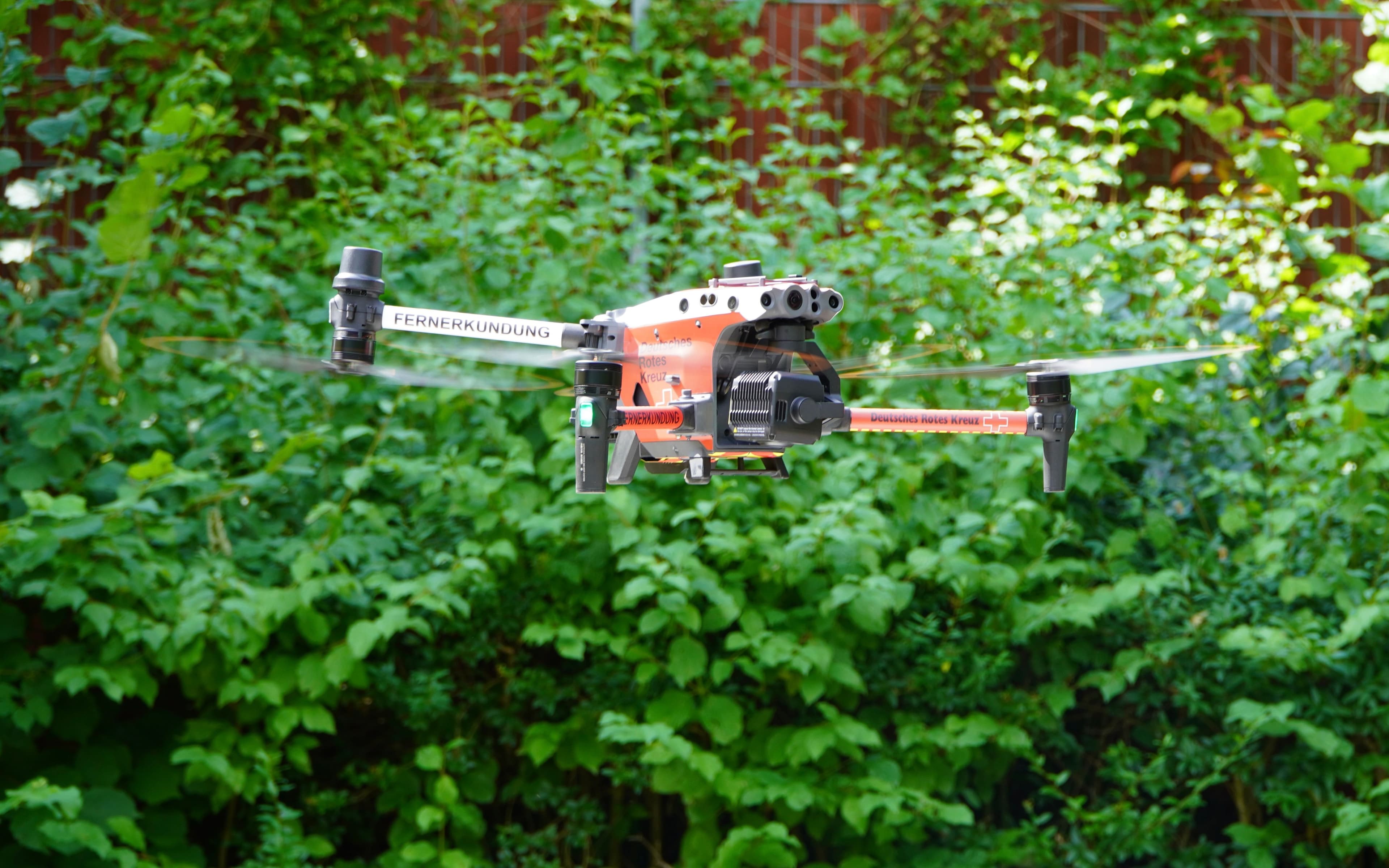 An orange and black drone with "FERNERKUNDUNG" written on its arm, hovering in front of green bushes.